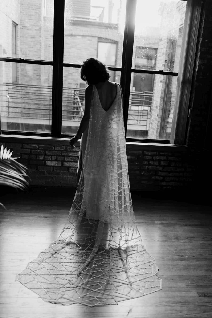 Bruid in NYC loft kijkend uit raam in trouw cape