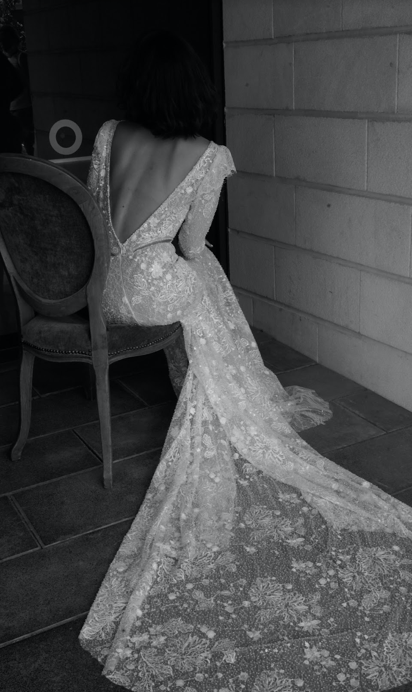 bruid op stoel met rug naar fotograaf in trouwjurk van kant
