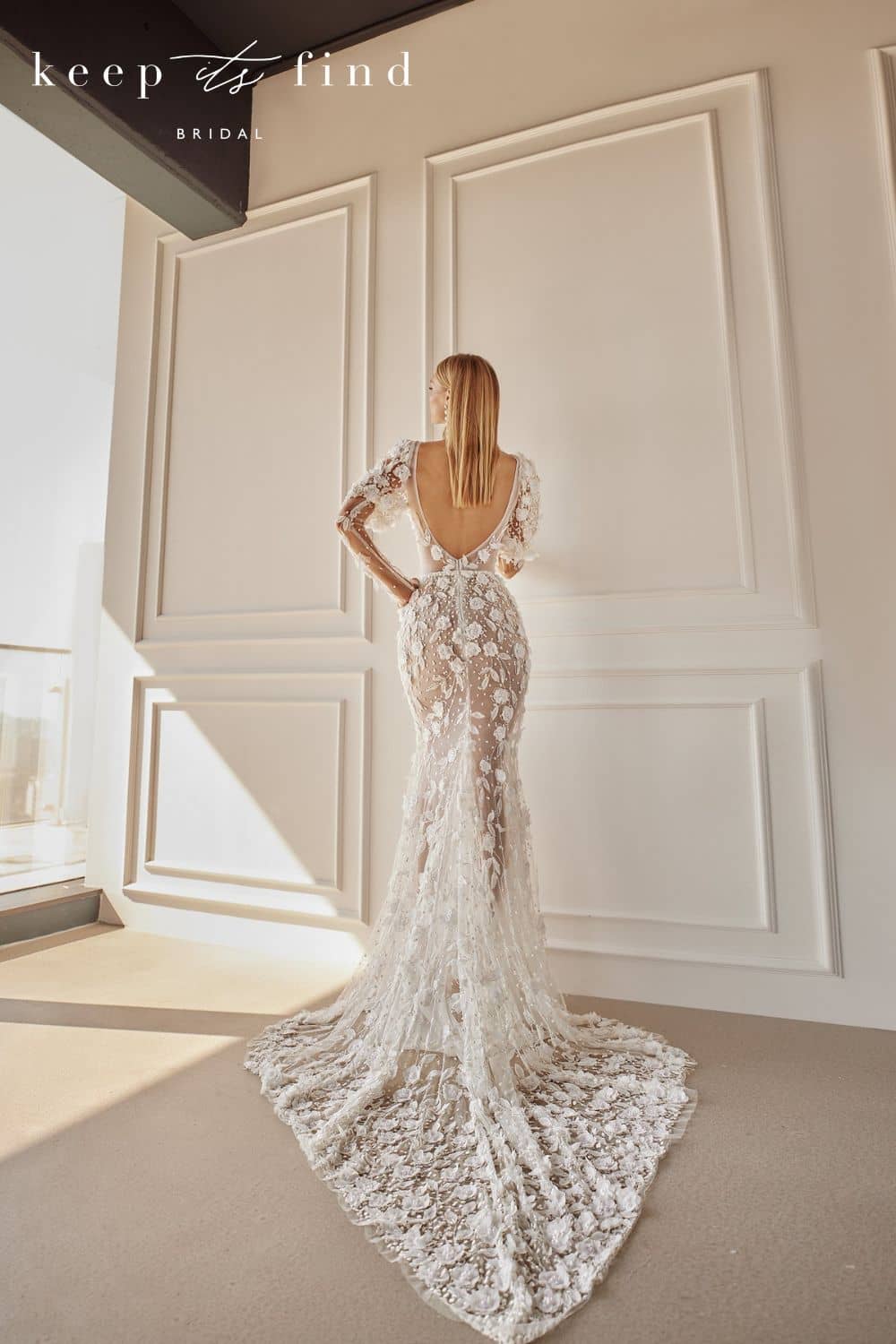 Bride standing in room with back toward camera in lace wedding dress with open back, long train and long sleeves