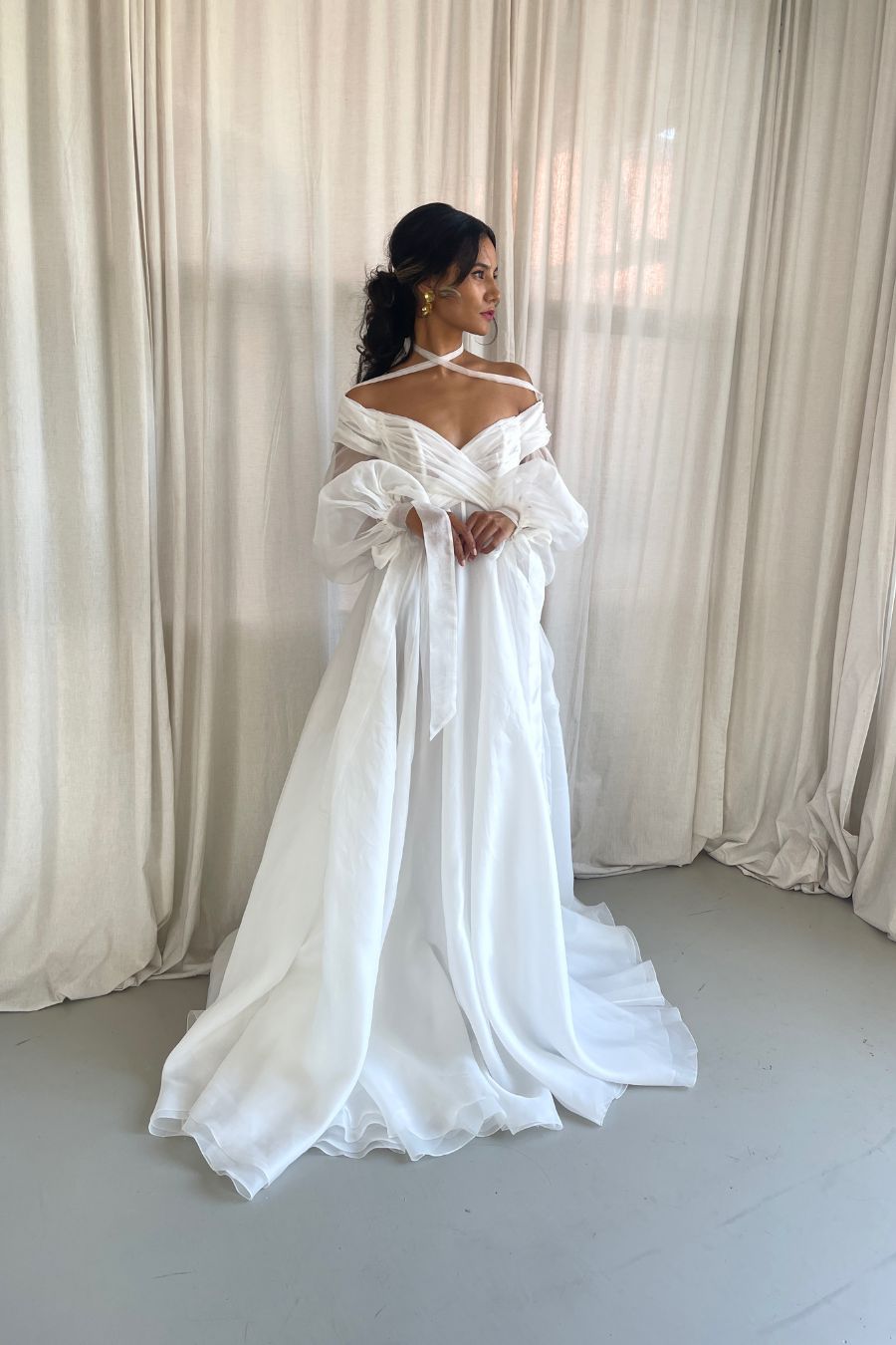 Bride standing with hands folded in aline silk wedding dress with ling sleeves and off shoulder neckline